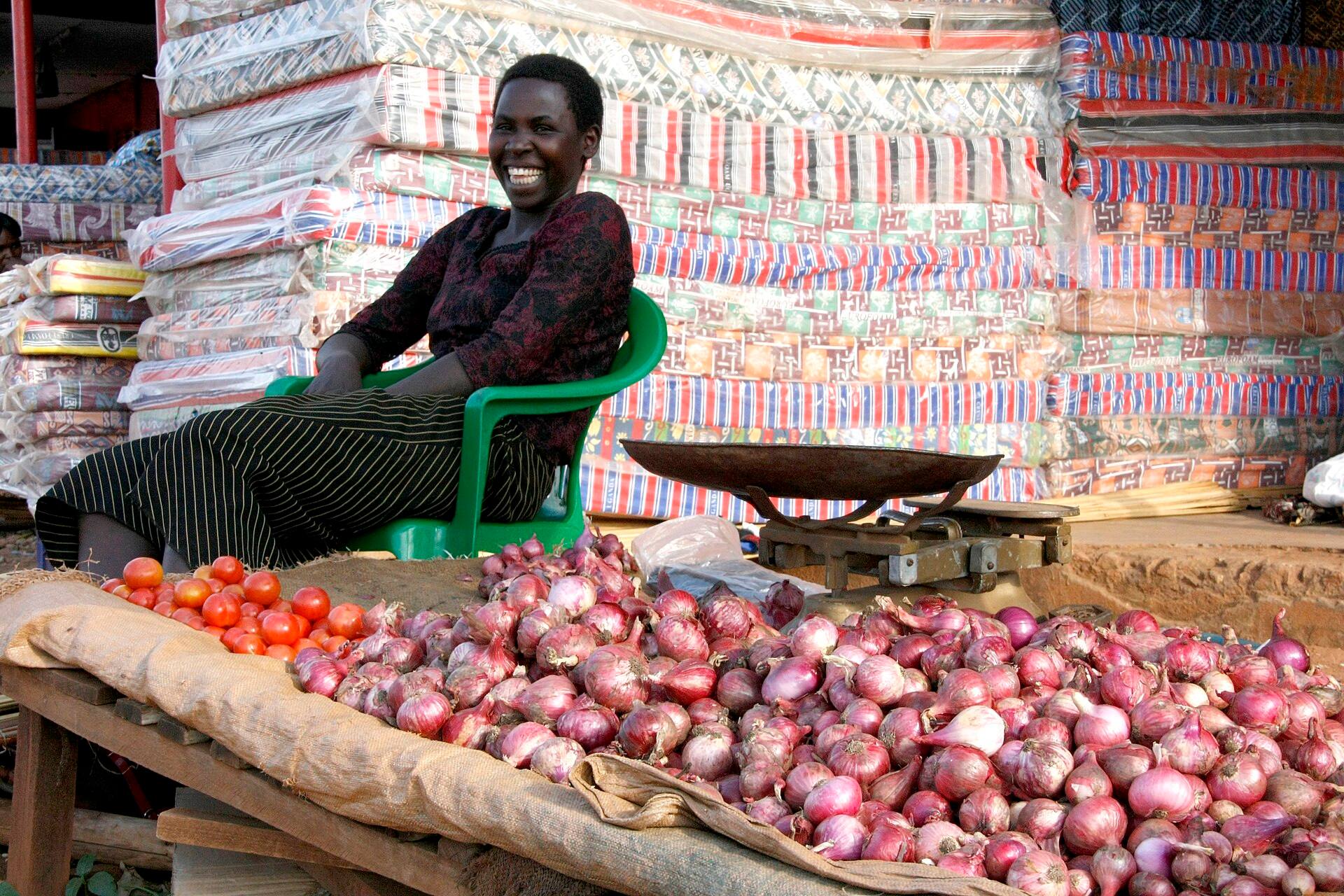 A smiling woman seated behind piles of red onions and tomatoes at her market stall