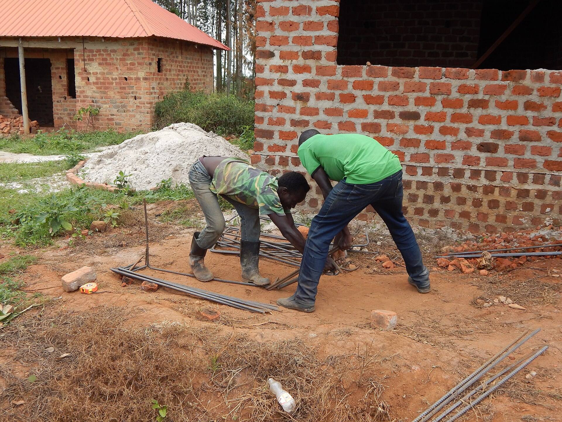 Two builders in green shirts bending steel reinforcing bars beside a brick wall